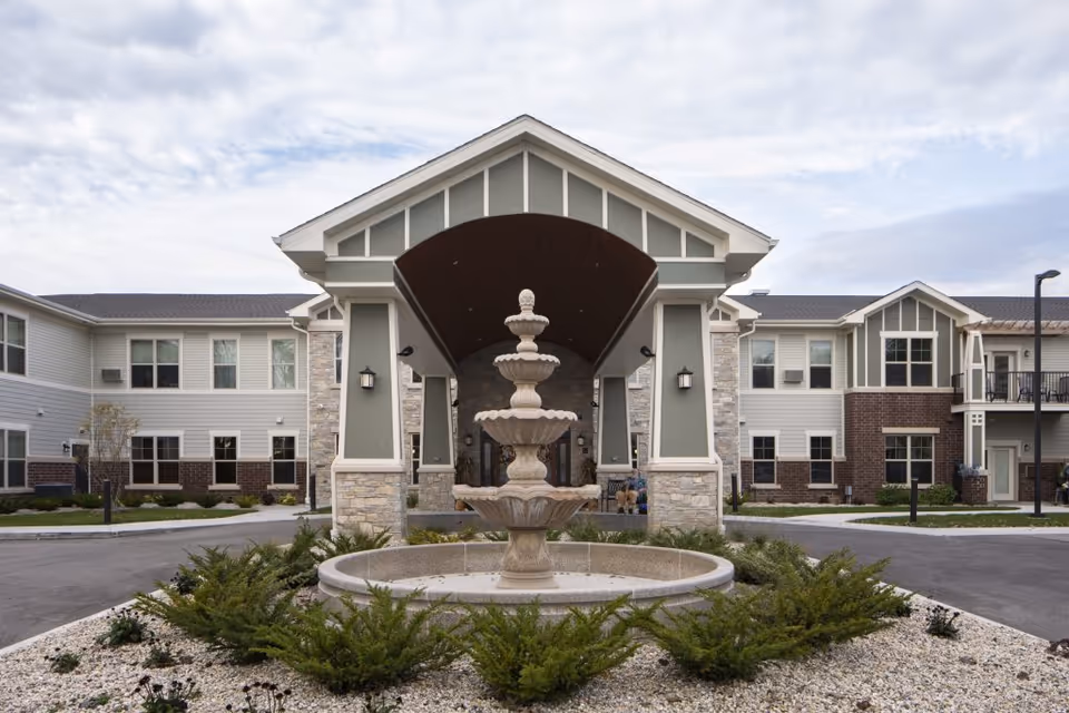 Front exterior view of Heritage Senior Campus featuring a large covered entrance with a multi-tiered stone fountain in the center surrounded by landscaping and shrubs. The building has two stories with a combination of stone, brick, and siding materials under a cloudy sky.