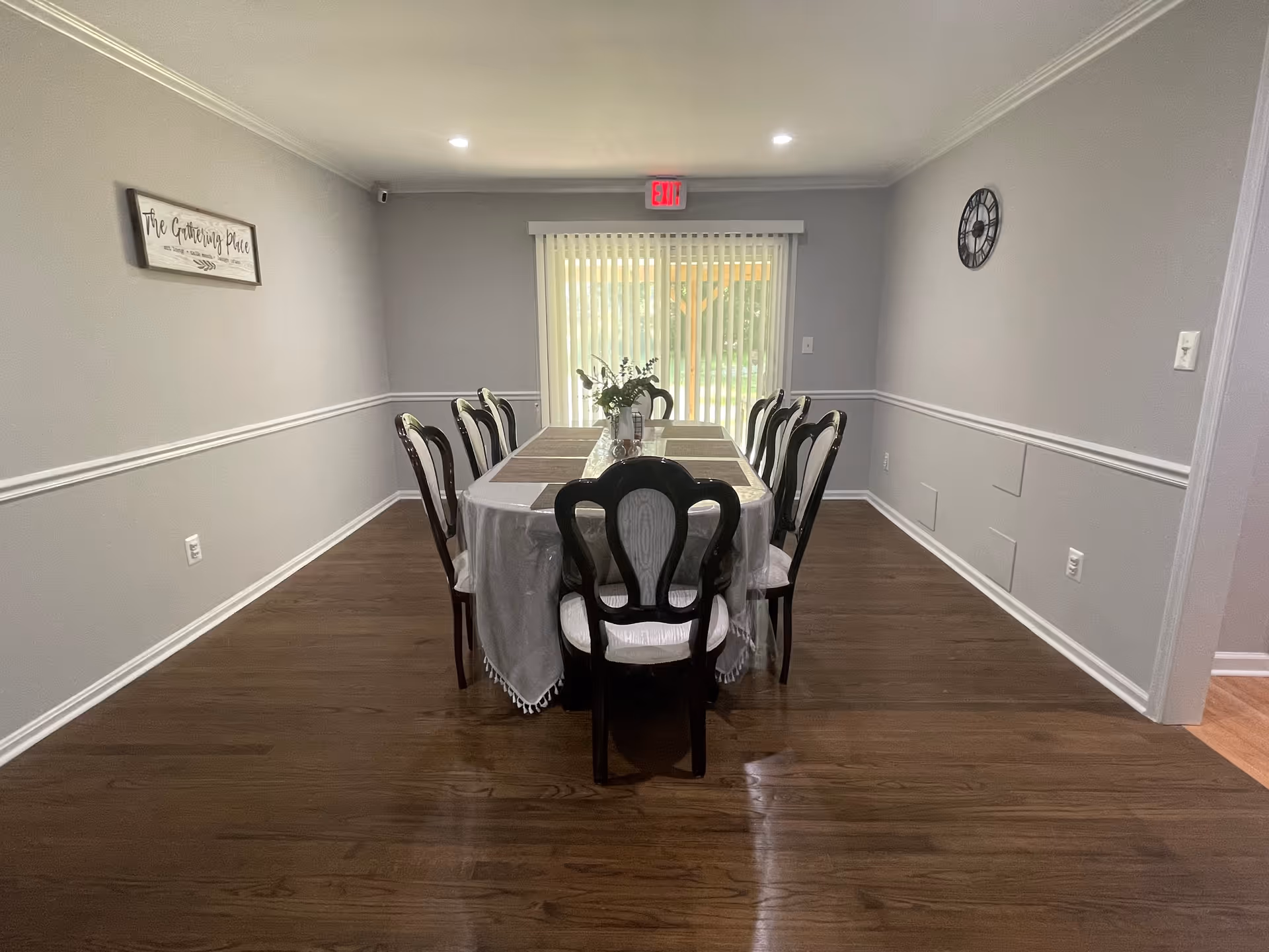 A dining room with a long rectangular table covered with a white tablecloth and placemats, surrounded by eight dark wooden chairs with white cushions. A vase with flowers is placed in the center of the table. The room has light gray walls with white trim, a wall clock on the right wall, a sign reading 'The Gathering Place' on the left wall, and vertical blinds covering a sliding glass door at the far end. The floor is wooden, and there is an exit sign above the door.