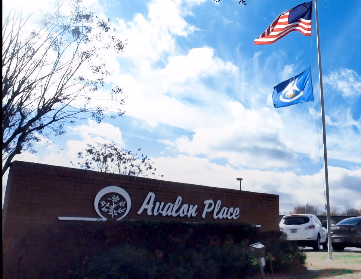 Outdoor view of a brick sign for Avalon Place with two flags flying on a flagpole against a partly cloudy blue sky. There are some trees and parked cars visible in the background.