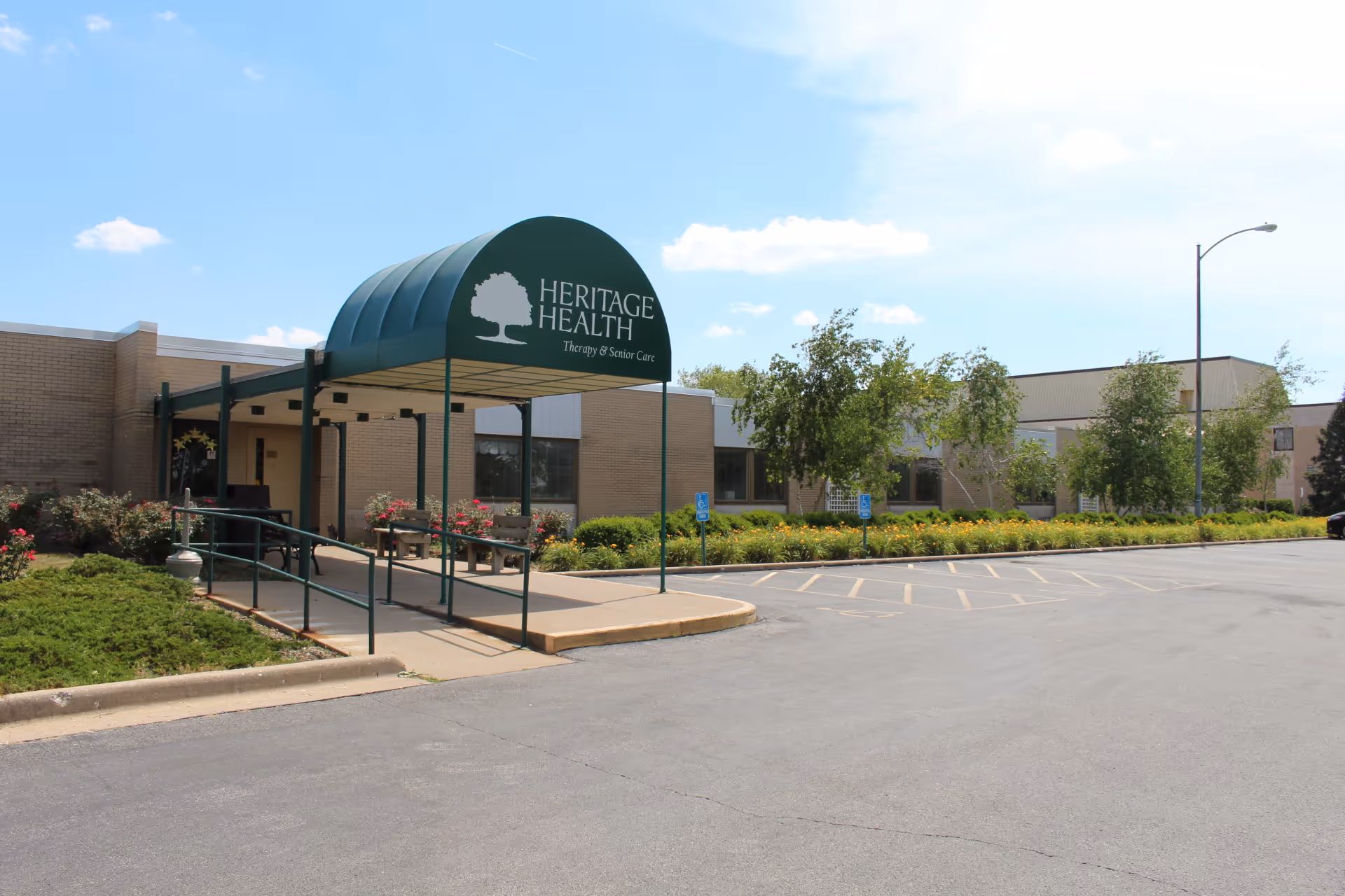 Front entrance of a single-story health center with a green canopy reading 'Heritage Health' and a nearby parking area and landscaping.