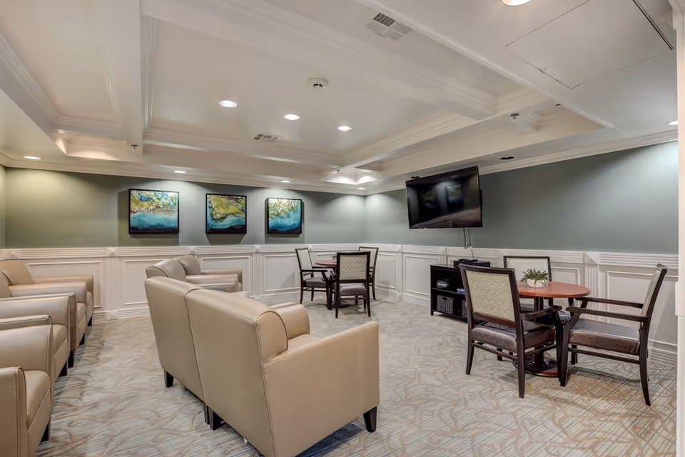 A cozy common area in Oakey Assisted Living featuring beige armchairs arranged in rows, two round tables each surrounded by four chairs, a wall-mounted flat screen TV, three abstract paintings on a green accent wall, and soft overhead lighting.