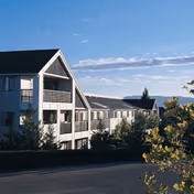 Exterior view of a multi-story residential building with balconies, surrounded by trees and bushes under a partly cloudy sky.