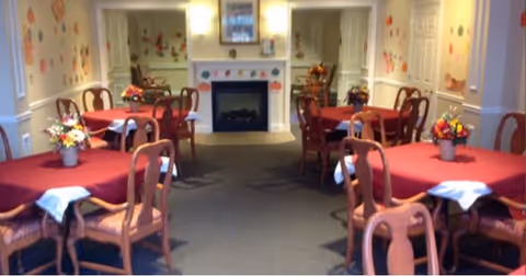 A dining room with several wooden tables covered with red tablecloths and white napkins. Each table has a floral centerpiece. The room features a fireplace at the far end with decorative artwork above it and floral wall decorations on the side walls.
