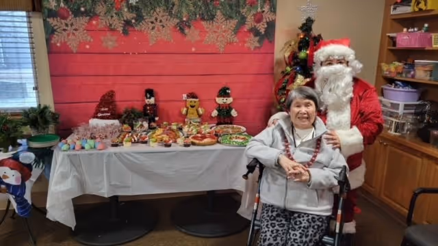 An elderly woman in a wheelchair smiling with a person dressed as Santa Claus standing behind her. They are indoors next to a table covered with a white tablecloth displaying various holiday-themed treats and decorations. The background features a red wall with gold snowflake decorations and a decorated Christmas tree.