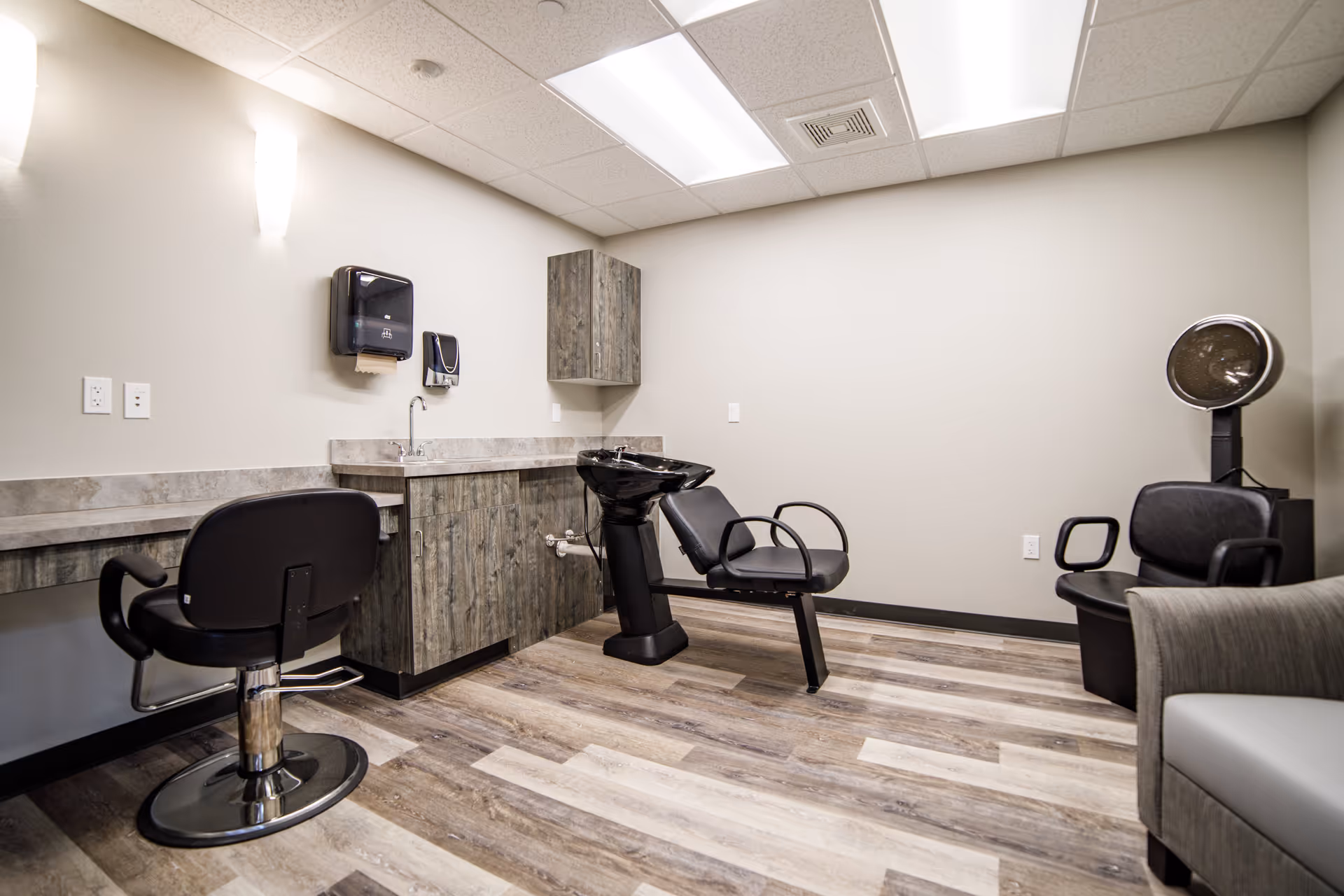 Interior view of a hair salon room in a senior living facility featuring a black salon chair in front of a counter with a sink, a black shampoo station chair with a basin, a black hair dryer chair, and a gray armchair. The room has wood-patterned flooring, light-colored walls, and ceiling lights.