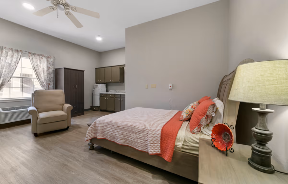 A senior living facility bedroom with a bed covered in a pink and red quilt, decorative pillows, a wooden nightstand with a lamp and a red decorative plate, a beige armchair near a window with floral curtains, a dark wooden wardrobe, and a kitchenette area with cabinets, a microwave, and a mini fridge.