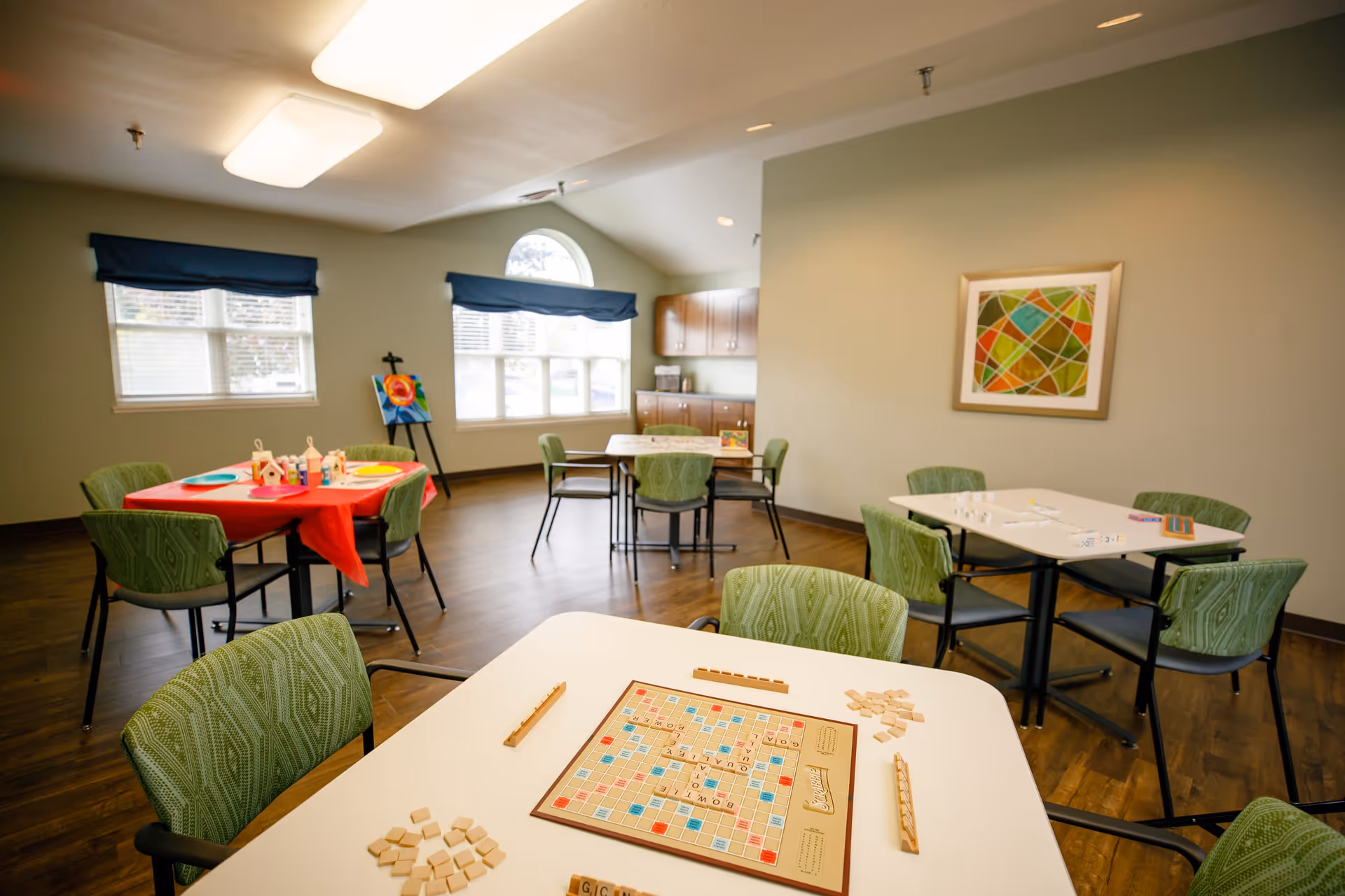 A well-lit activity room with multiple tables and green cushioned chairs. One table in the foreground has a Scrabble board game set up, while other tables have various games and craft supplies. The room has large windows with blue valances, wooden flooring, and a colorful abstract painting on the wall.