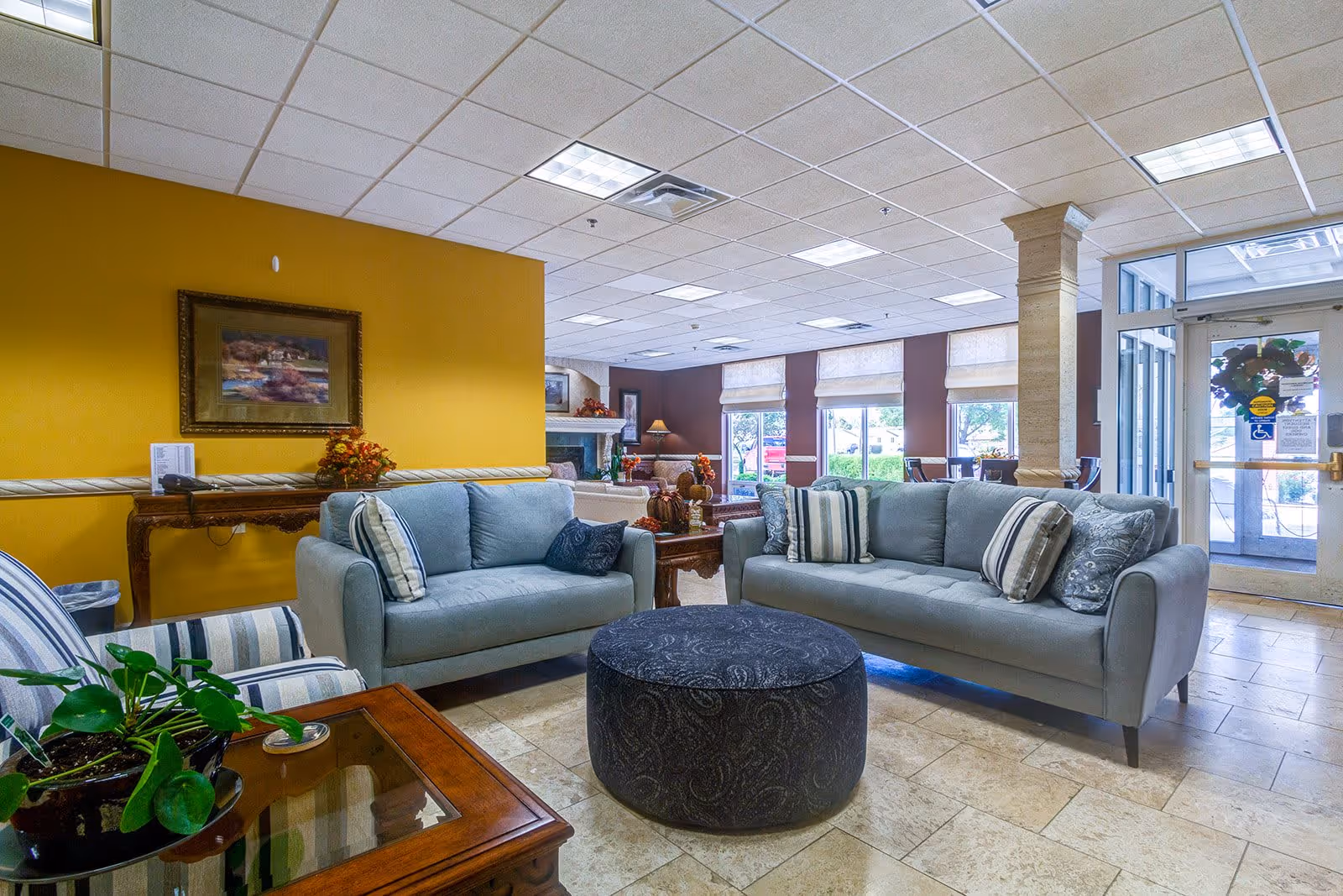 A bright and welcoming senior living facility common area with two light blue sofas arranged around a round dark ottoman. The room features a yellow accent wall with a framed painting, a wooden table with a flower arrangement, and large windows letting in natural light. There is a glass door entrance visible on the right side.