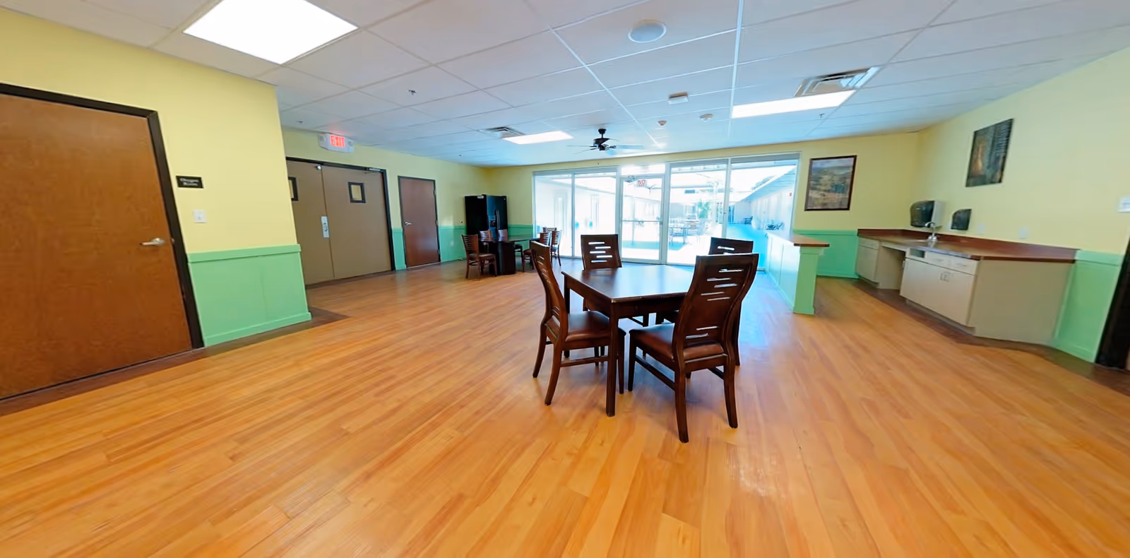 A spacious room with wooden flooring and light yellow walls with green wainscoting. There are two wooden tables with chairs arranged around them. The room has a ceiling fan, fluorescent lights, and large glass sliding doors leading to an outdoor patio area. On the right side, there is a counter with cabinets, a sink, and two wall-mounted soap or sanitizer dispensers. Several doors are visible on the left wall.