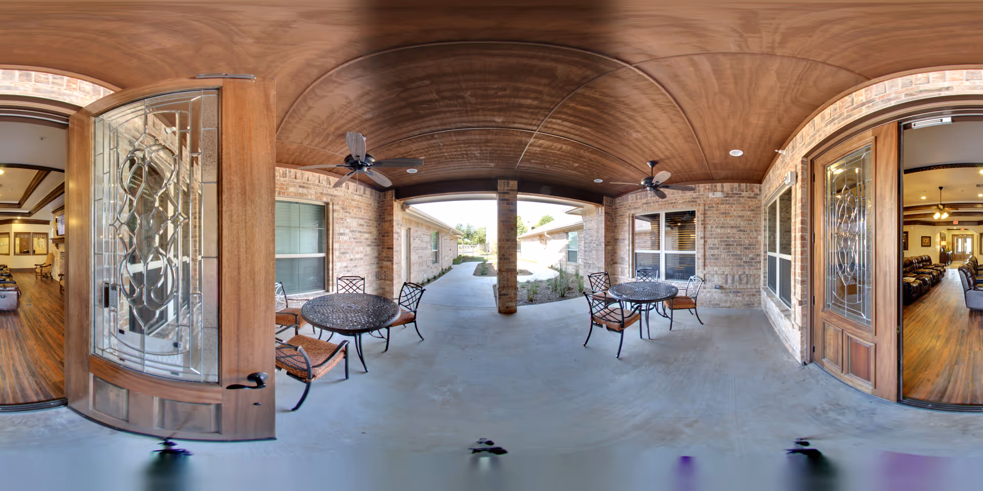 Covered outdoor patio area with a wooden ceiling and ceiling fans, featuring two round metal tables each surrounded by four chairs with woven seats. The patio is flanked by brick walls with windows and glass doors leading into an interior space with wooden floors and furniture visible inside.