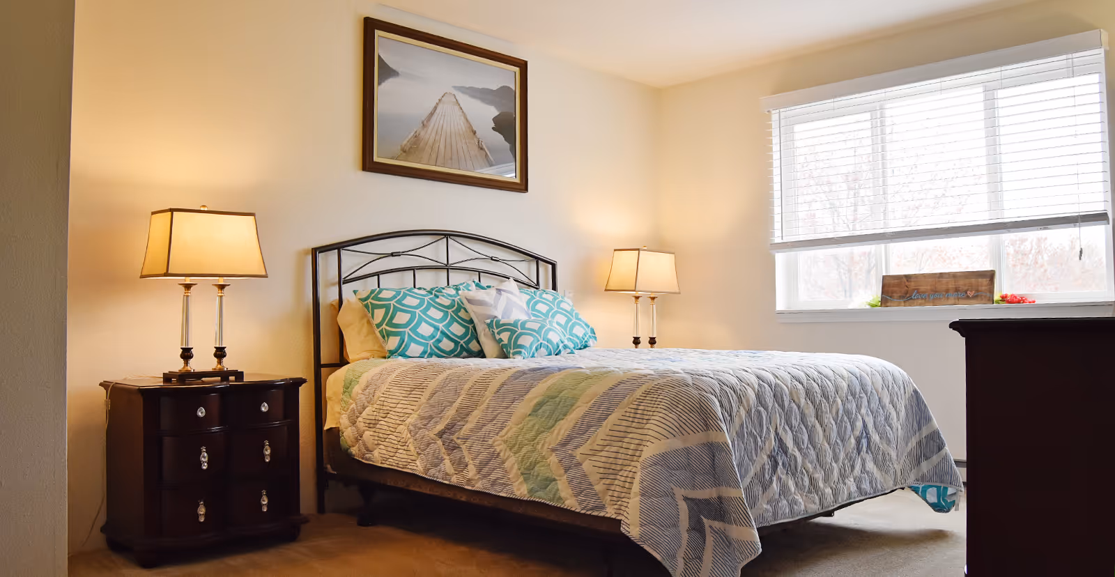 A bright bedroom with a double bed, patterned pillows and quilt, two bedside tables with lamps, a framed picture above the headboard, and a window with blinds.