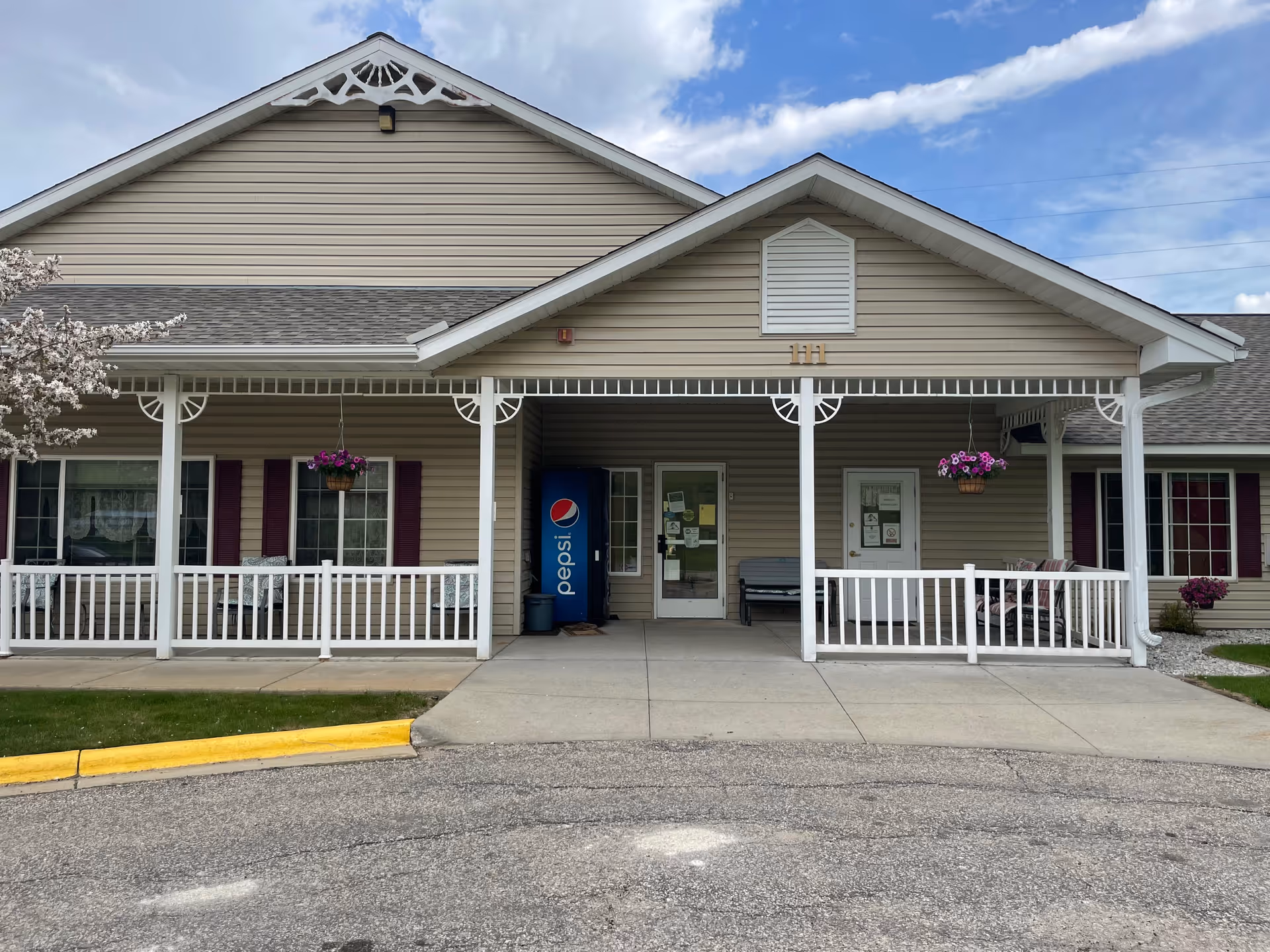 Front exterior view of a single-story building with beige siding and a covered porch. The porch has white railings, hanging flower baskets, and seating areas. There is a Pepsi vending machine near the entrance door. The building number 111 is displayed above the porch.