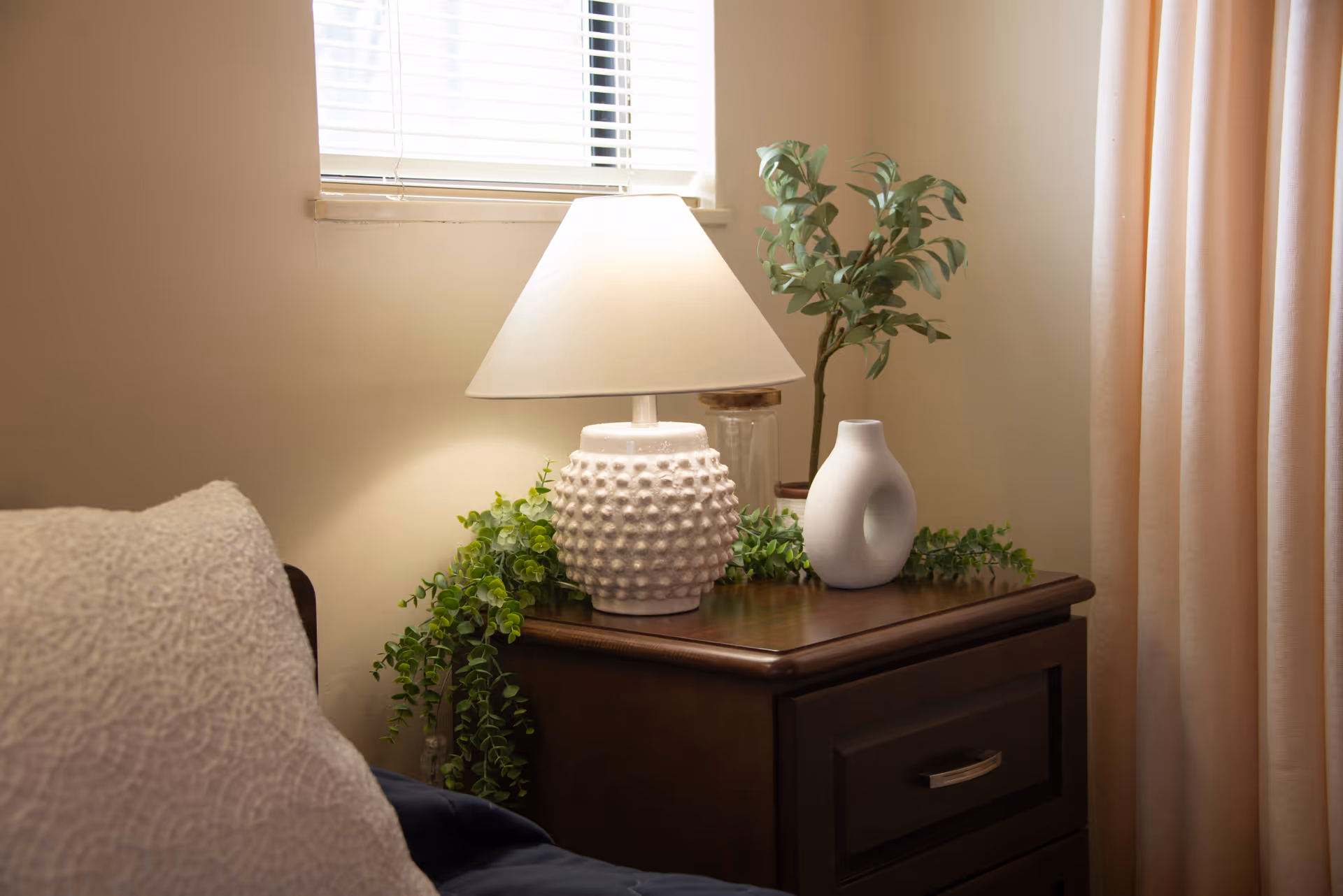 A bedside table in a bedroom holds a textured lamp, decorative vases, and greenery next to a bed and window.