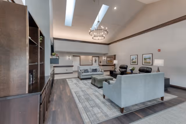 A spacious senior living facility common area with high ceilings and skylights. The room features a large area rug with two gray sofas, two black armchairs, and a central ottoman. There is a dark wood built-in shelving unit on the left side and framed artwork on the walls. The space is well-lit with ceiling lights and table lamps.