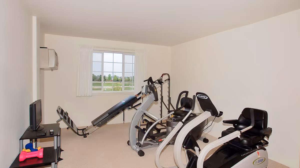 Small exercise room with multiple cardio machines, a TV on a small stand, and a window.