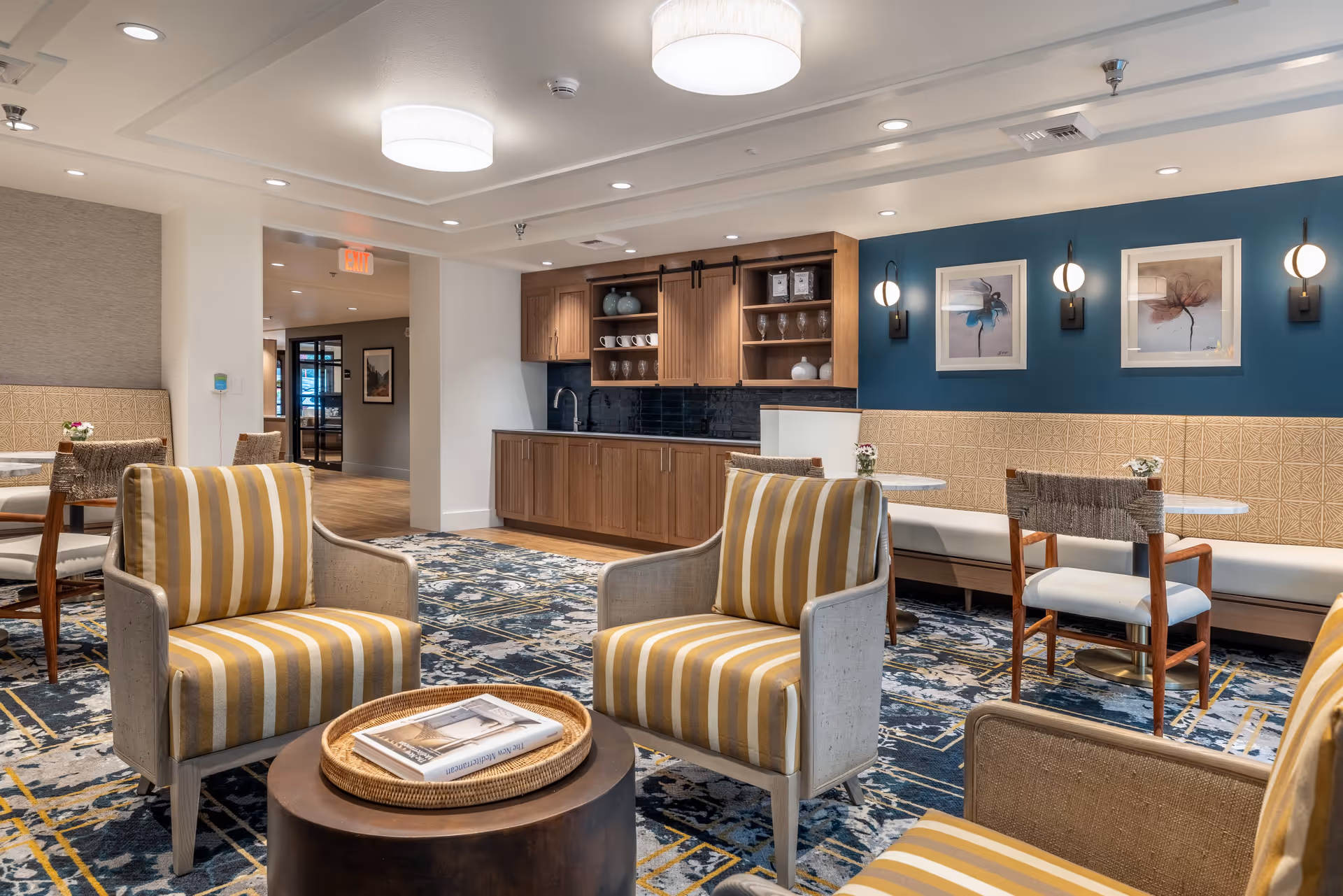 A cozy senior living common area with striped armchairs around a small round table, a kitchenette with wooden cabinets and a sink, and a seating area with tables and chairs against a blue accent wall decorated with framed artwork and wall sconces.