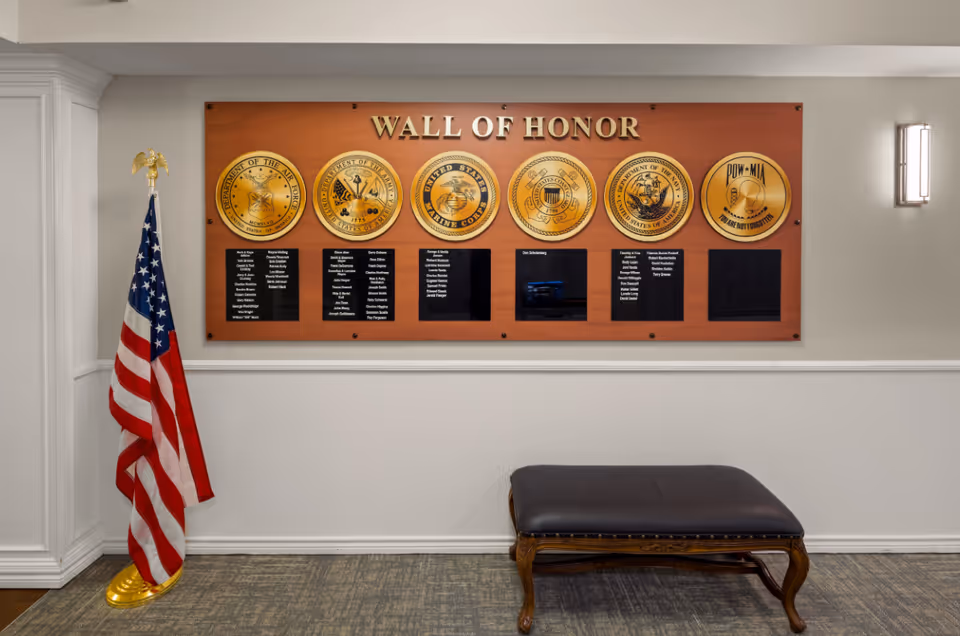 An indoor wall display titled 'Wall of Honor' featuring emblems of various U.S. military branches including the Air Force, Navy, Marine Corps, Coast Guard, Army, and a POW-MIA emblem. Below each emblem are black plaques with names inscribed. To the left of the display is a standing American flag with a golden eagle finial. In front of the wall is a dark cushioned bench with wooden legs. The room has neutral-colored walls and carpeted flooring.