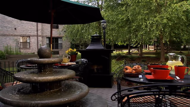 Outdoor courtyard patio with a tiered fountain, metal tables set with red cups, pastries and a pitcher, and a small outdoor fireplace beneath trees.