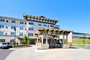 Front exterior of a multi-story senior living building with a covered porte-cochere entrance, driveway, and parked cars under a clear blue sky.