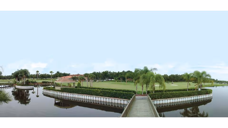 A panoramic outdoor view of a landscaped area with a wooden walkway leading to a green space surrounded by water. The area features palm trees, manicured bushes, and a grassy field with a few people in the distance. The sky is clear and blue.