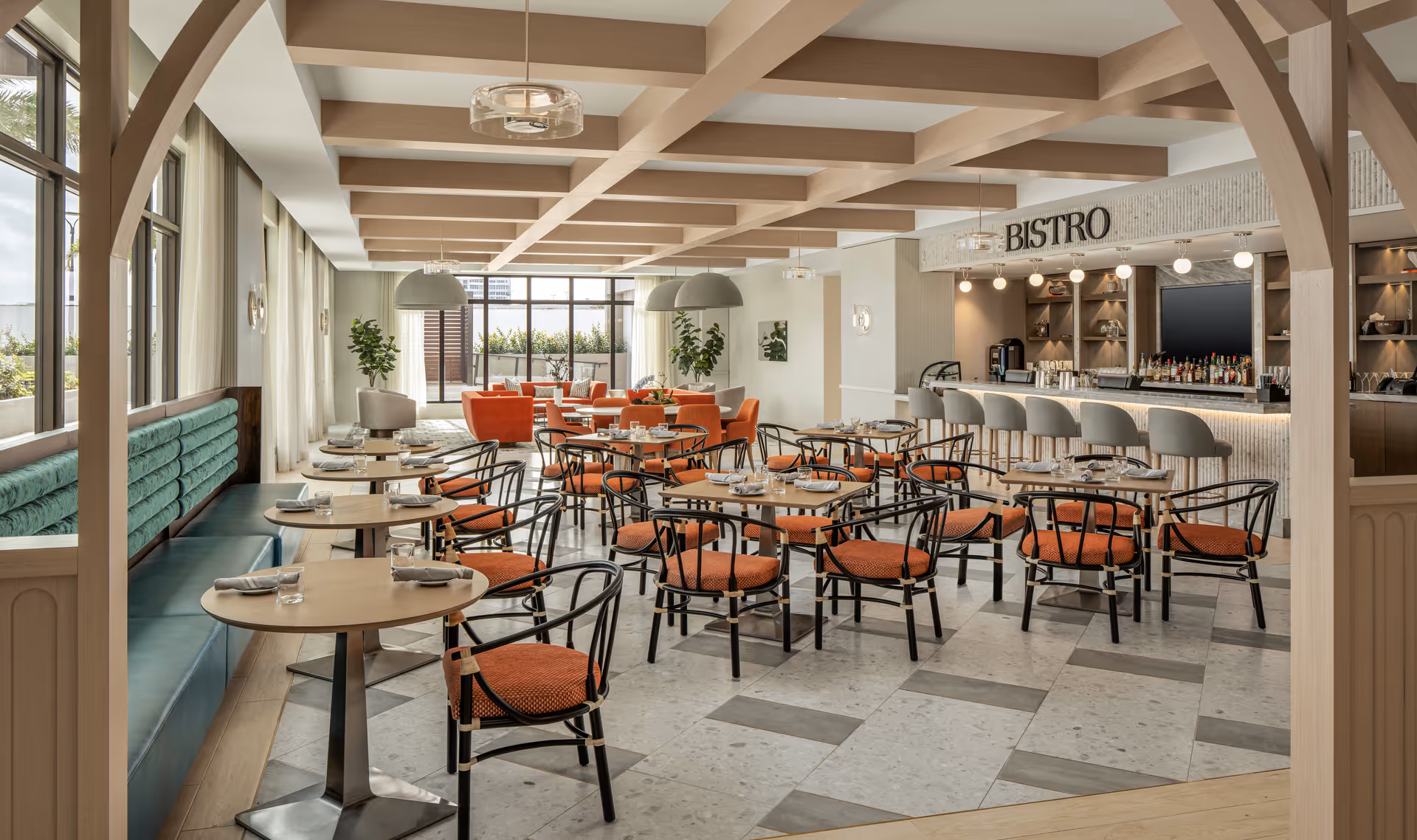 A spacious and well-lit dining area with multiple round tables set with napkins and glasses. The chairs have orange cushions and black frames. There is a long teal cushioned bench along one side and a bar area labeled 'BISTRO' with bar stools on the opposite side. Large windows allow natural light to fill the room, and there are plants and modern light fixtures adding to the decor.