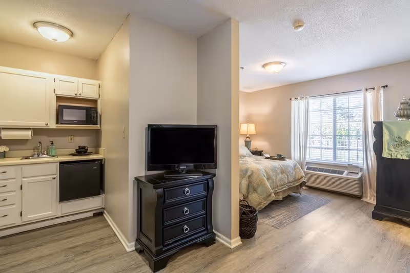Interior view of a senior living facility room showing a small kitchen area with white cabinets, a microwave, and a mini fridge on the left. A black TV stand with a flat-screen TV is positioned against a partial wall dividing the kitchen from the bedroom area. The bedroom area has a bed with a patterned comforter, a nightstand with a lamp, a large window with white curtains, and an air conditioning unit below the window.