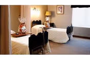 A room with two single beds, each with white bedding and a black headboard. There is a bedside table between the beds with a lamp and a decorative item. A window with closed blinds is visible on the right side of the room, and a framed picture hangs on the wall above the lamp.