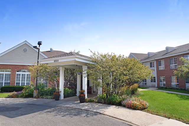 Exterior view of Eden Vista Burr Ridge senior living facility showing a covered entrance with the number 6801, surrounded by landscaped greenery, flowering plants, and a well-maintained lawn under a clear blue sky.