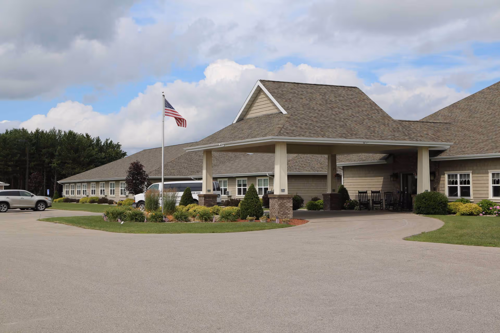 Exterior view of The Brook of Gaylord facility showing a single-story building with a covered entrance supported by columns. There is an American flag on a flagpole in front of the building, surrounded by landscaped greenery and flowers. Several vehicles are parked near the entrance under a partly cloudy sky.