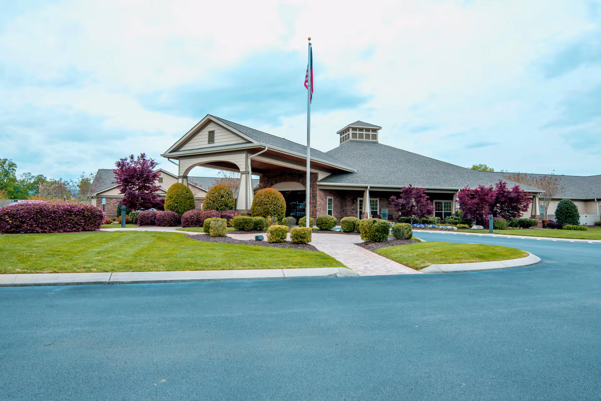 Front entrance of a senior living building with a covered porte-cochère, flagpole, and landscaped lawn.