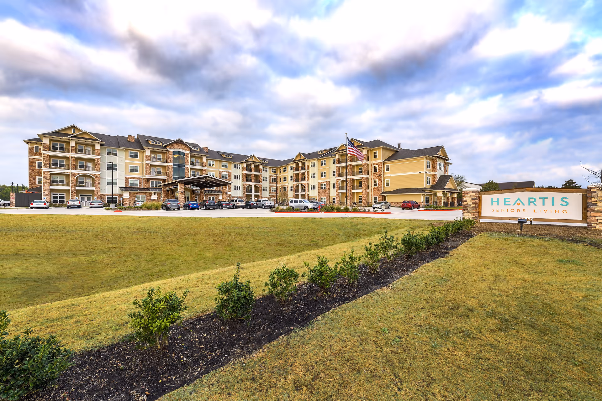 Exterior view of a large senior living facility building with multiple floors, a covered entrance, parked cars, and a landscaped lawn with small bushes. A sign in front reads 'Heartis Seniors Living.' The sky is partly cloudy.