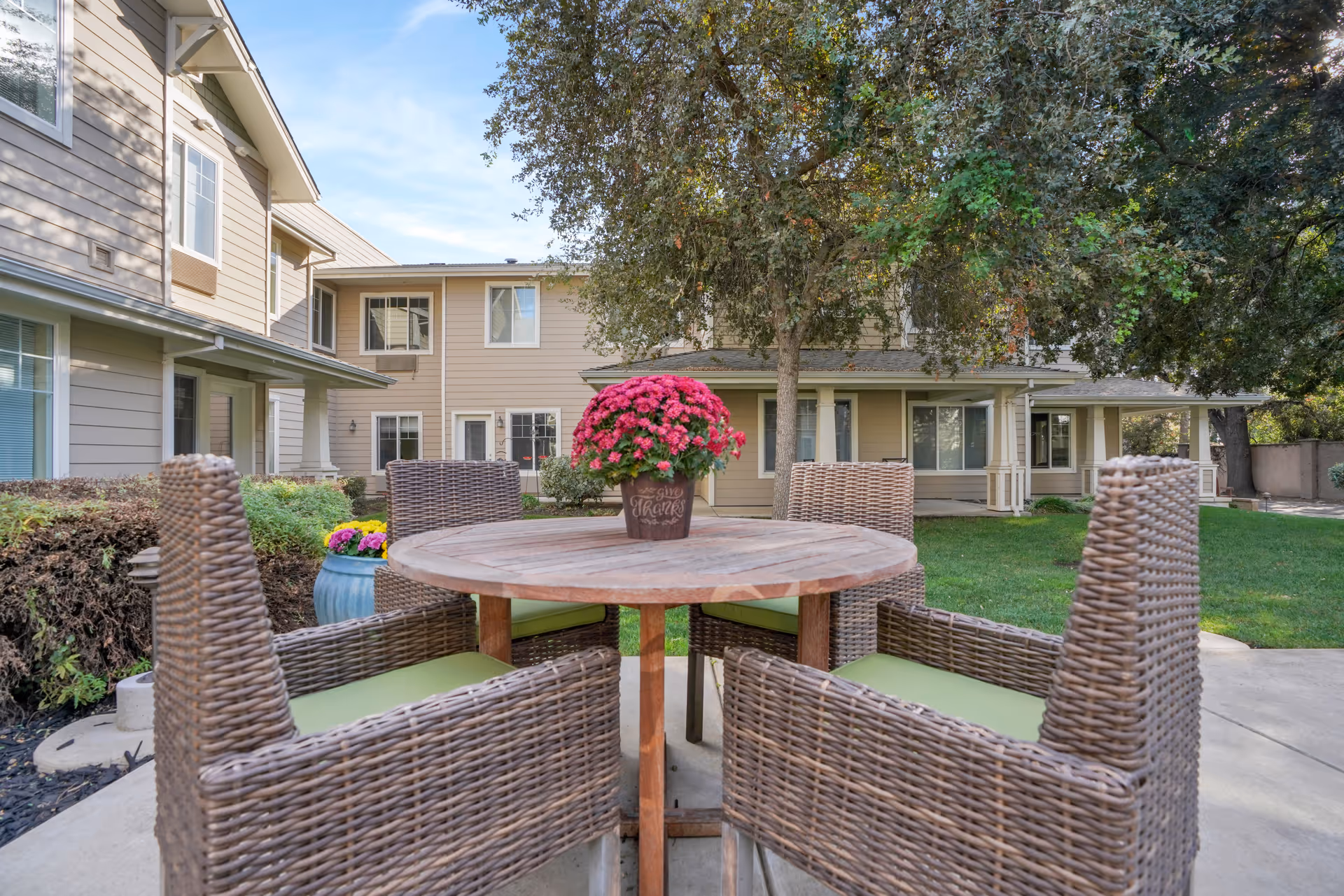 Outdoor patio area at The Commons At Elk Grove featuring a round wooden table with a potted plant with pink flowers in the center, surrounded by four wicker chairs with green cushions. The background shows a two-story beige building with multiple windows and a well-maintained lawn with trees and shrubs.