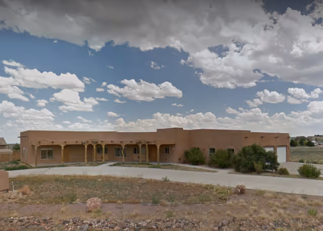 Single-story adobe-style building with a covered porch, driveway, and dry landscaping under a partly cloudy sky.