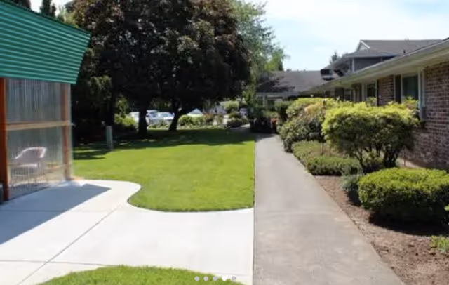Outdoor pathway alongside a building with bushes and trees, a green lawn, and a covered seating area with a chair on the left side.