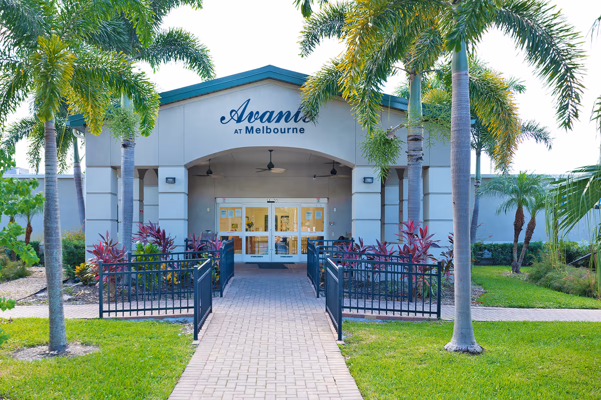 Front entrance of Avante at Melbourne senior living facility framed by palm trees and a paved walkway.