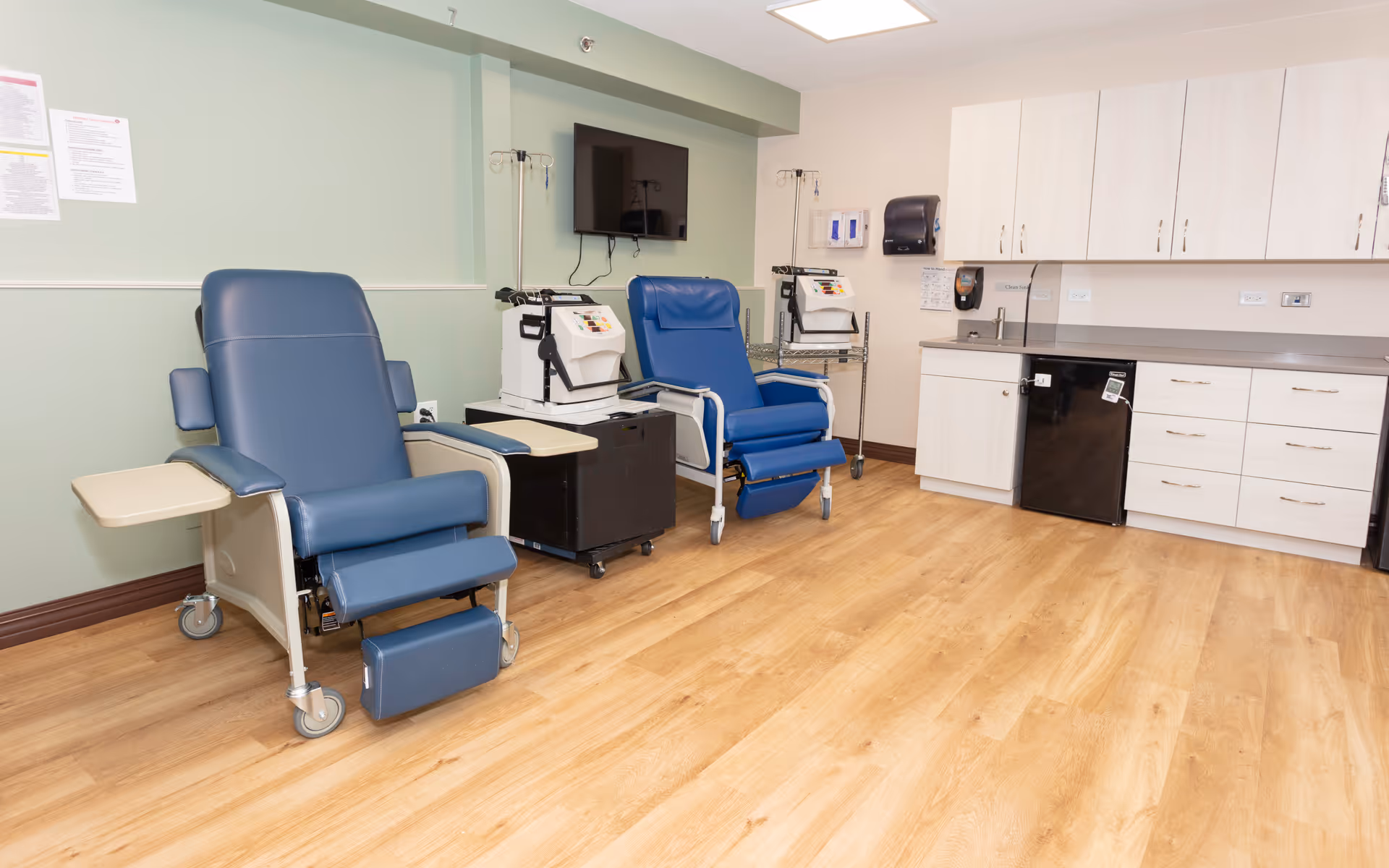 A medical treatment room with two blue reclining chairs on wheels, each equipped with armrests and small attached tables. Behind the chairs are medical devices on carts, and a wall-mounted TV is visible. The room has light green and beige walls, wooden flooring, and white cabinets with a countertop and a small black refrigerator.