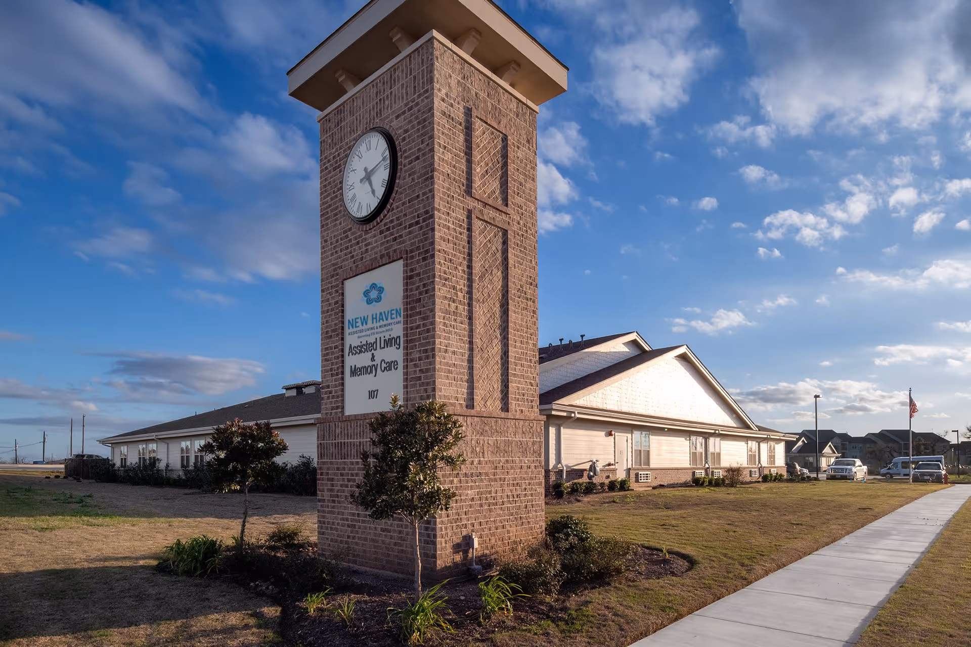 Exterior view of New Haven Assisted Living and Memory Care of Floresville building with a prominent brick clock tower displaying the facility's name and logo. The building is surrounded by a lawn, small trees, and a sidewalk leading to the entrance under a partly cloudy sky.