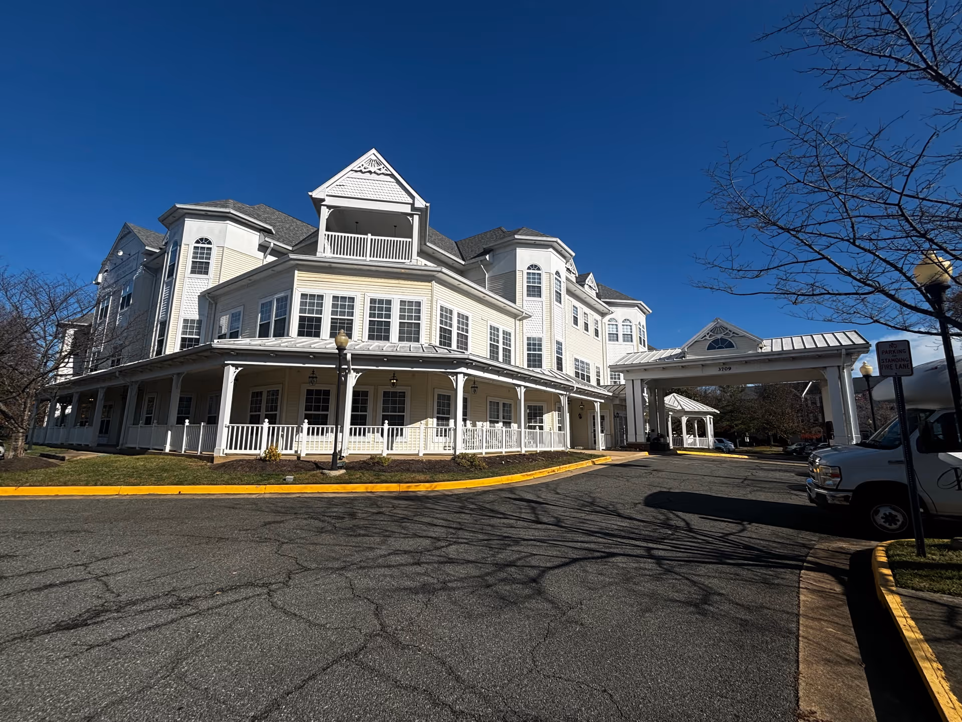 Exterior view of a large, multi-story senior living facility building with white siding and multiple windows under a clear blue sky. The building has a covered entrance driveway with a white portico and a wrap-around porch with white railings. There are leafless trees and a paved driveway in front.