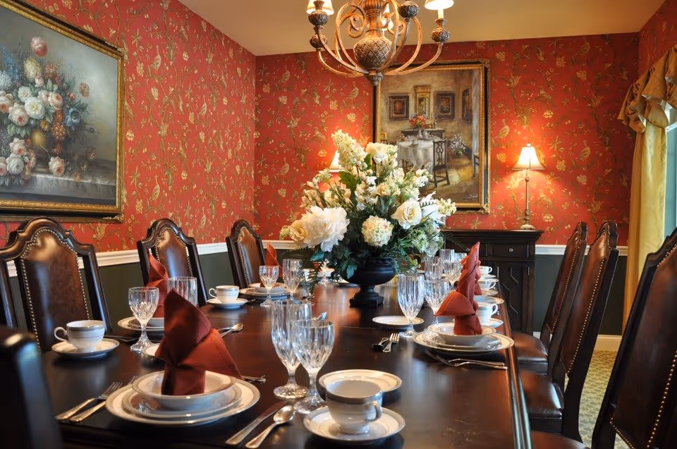 Elegantly set dining room with a long polished table, leather chairs, crystal glassware, a large floral centerpiece, chandelier, and red patterned wallpaper.