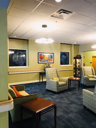 A cozy common area with several cushioned armchairs arranged around small wooden tables. The walls are painted light green and decorated with framed artwork and a glass display cabinet. The ceiling has recessed lighting and a hanging light fixture.