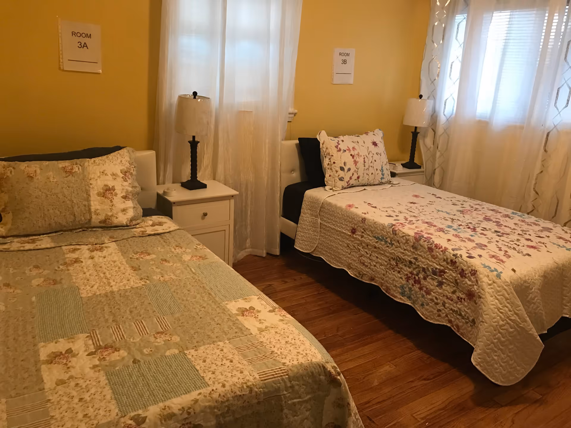A cozy bedroom with two single beds, each with floral-patterned bedspreads and pillows. Between the beds is a white nightstand with a lamp on top. The walls are painted yellow, and there are sheer white curtains on the windows behind the beds. Signs above each bed read 'ROOM 3A' and 'ROOM 3B'.