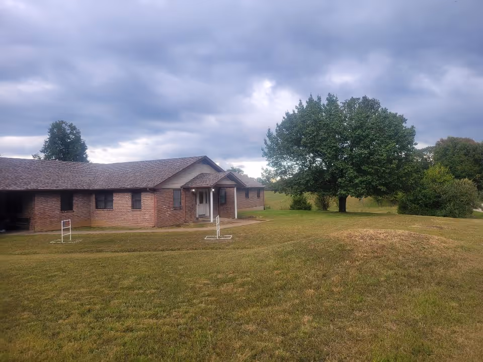 Single-story brick building with a covered front entrance, grassy lawn and a large tree beneath a cloudy sky.