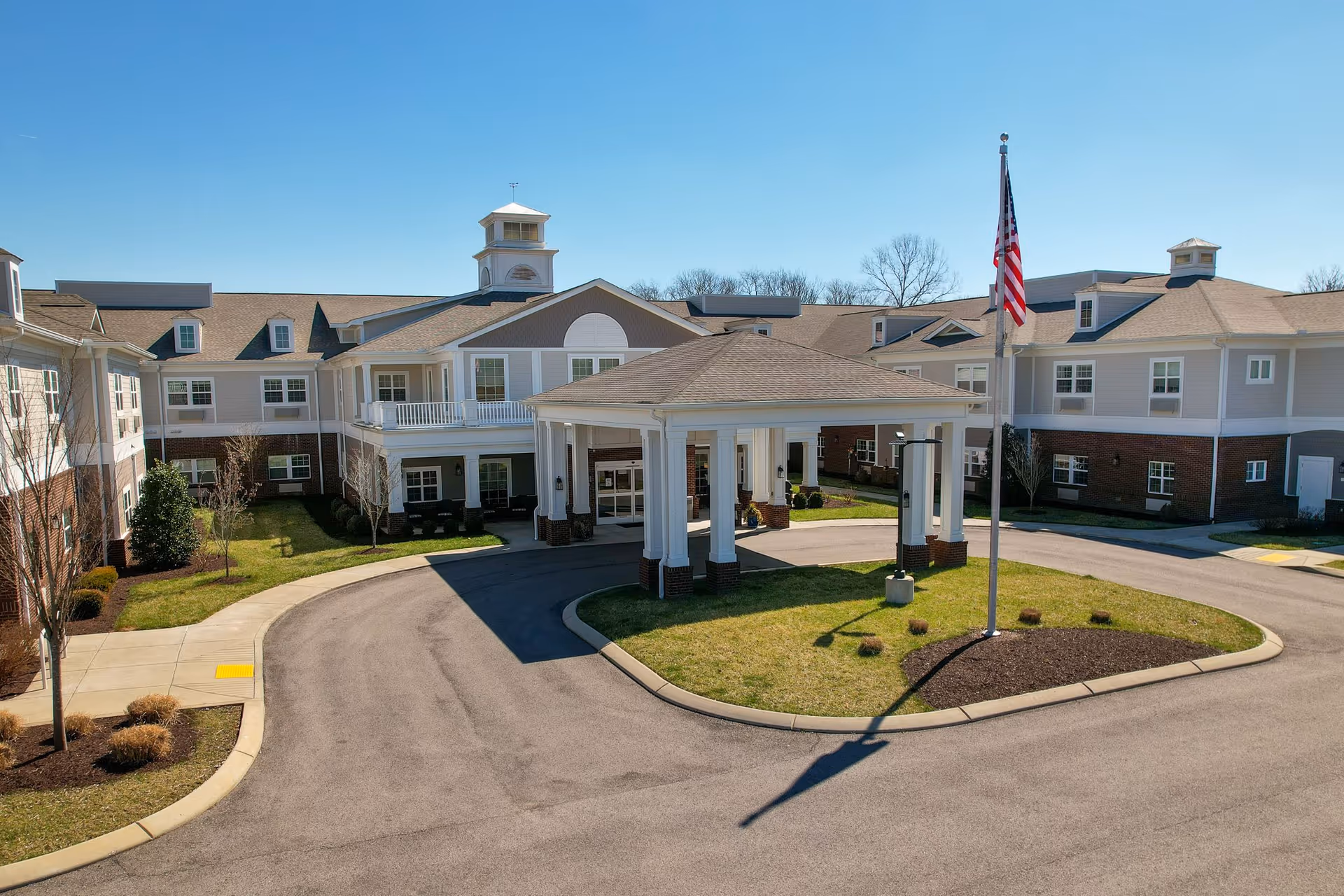 Exterior view of The Goldton at Spring Hill senior living facility showing a large, two-story building with a covered entrance driveway, an American flag on a flagpole, and landscaped grounds under a clear blue sky.
