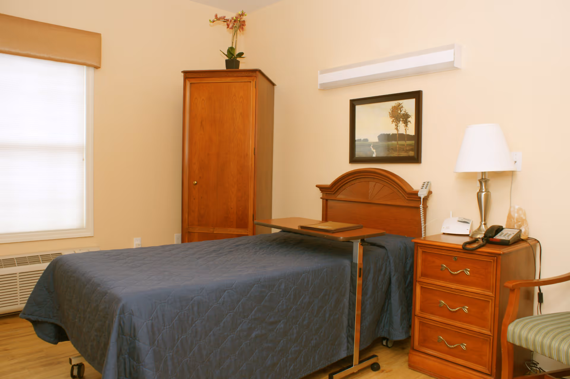A bedroom in a senior living facility with a bed covered in a dark blue quilt, a wooden headboard, a wooden nightstand with a lamp, telephone, and other devices, a wooden wardrobe, a window with a beige shade, and a framed landscape painting on the wall.