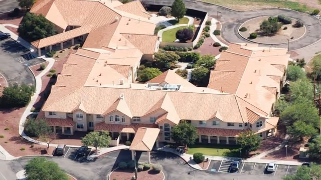 Aerial view of a large senior living facility with a U-shaped building featuring a tiled roof, surrounded by landscaped greenery, parking areas, and a circular driveway.