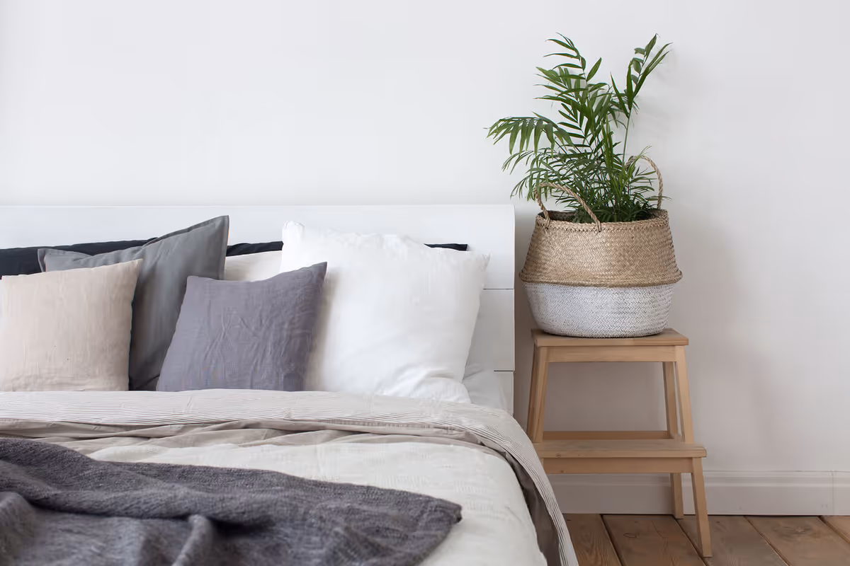 A neatly made bed with several pillows beside a wooden stool holding a woven basket with a green plant.
