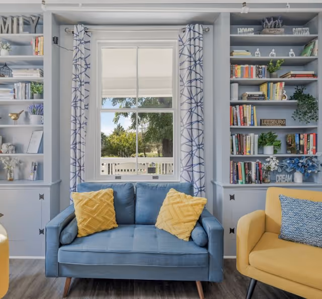 Bright living room with a blue loveseat in front of a window, built-in bookshelves on either side, and a yellow armchair at right.