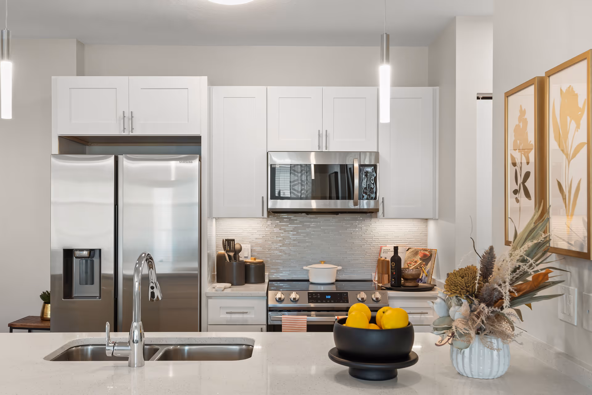 Modern kitchen with white cabinetry, stainless steel refrigerator and microwave, a stove with a white pot on it, a double sink with a chrome faucet, a black bowl with yellow lemons on the counter, and a white vase with dried flowers. Two framed botanical prints hang on the wall to the right.