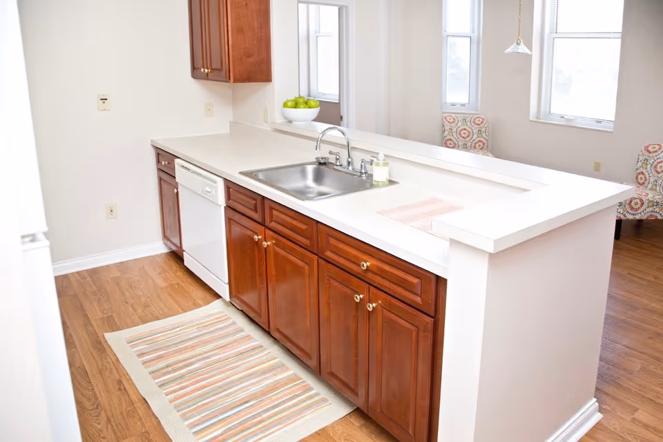 A bright kitchen area with wooden cabinets, a white countertop, a stainless steel sink with a faucet, a white dishwasher, and a striped rug on the wooden floor. In the background, there are windows with blinds and patterned chairs near a dining or living area.