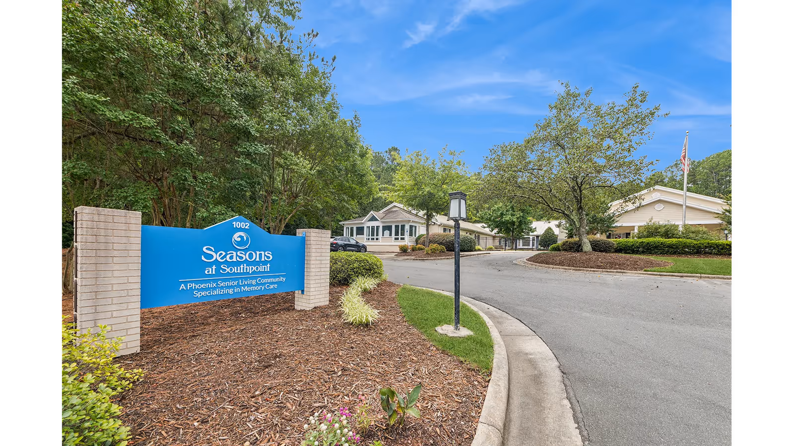 Entrance sign for Seasons at Southpoint, a senior living community specializing in memory care, with a driveway leading to single-story buildings surrounded by trees and landscaping under a blue sky.